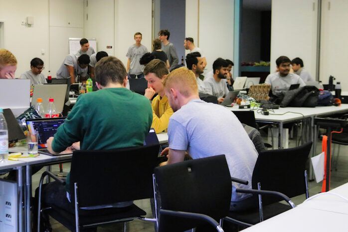 Viele Hackathon-Teilnehmer*innen sitzen in einem Raum an Tischen, blicken in Laptops und arbeiten intensiv.